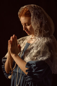 Caucasian young adult woman praying with hands pressed together, holding rosary beads, wearing lace veil over head, eyes closed, standing against dark background