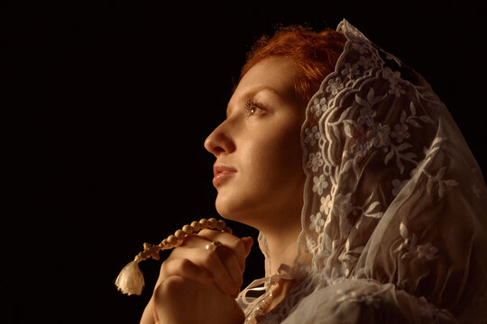 Caucasian young adult woman wearing lace veil holding prayer beads gazing upward in profile view showing serene facial expression with soft lighting highlighting facial features
