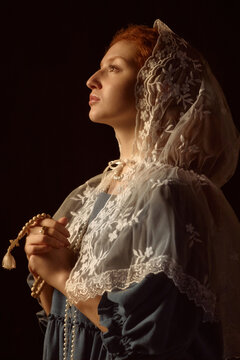Caucasian young adult woman wearing lace headscarf holding rosary beads praying with hands clasped looking upward side view religious devotion concept dark background