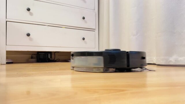 A modern robotic vacuum cleaner autonomously navigates across a hardwood floor, quietly cleaning the surface with steady, low hum of the vacuum motor. 