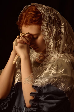 Caucasian young adult woman praying with rosary beads, wearing lace veil over head, eyes closed, hands clasped together, side profile visible against dark background