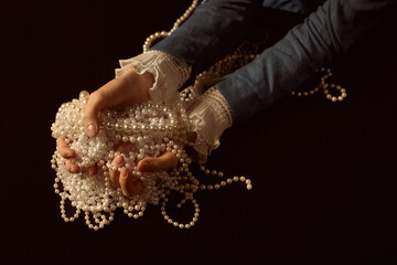 Caucasian young adult woman holding strands of pearls in both hands, wearing long sleeve garment with lace cuffs, displaying jewelry against dark background, hands gently cupping beads