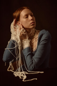 Portrait of young adult Caucasian woman closing eyes, holding multiple strands of pearls against face and neck, wearing lace collar and long sleeve garment, posing against dark background