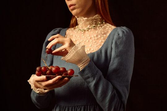 Caucasian young adult woman holding plate of red grapes, gently picking grape with right hand, wearing vintage style dress with lace collar, standing against dark background