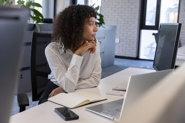African american woman resting chin on hand and gazing right at white desk in office with notebook