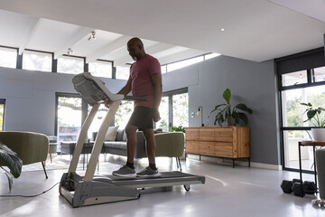Fototapeta premium Man running on treadmill in living room flooded by natural light with sofa, dumbbells, plants