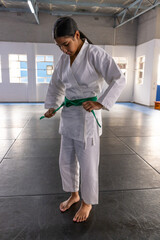Teenage asian girl standing on mats in martial hall tying green belt on white gi