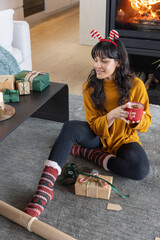 Naklejka premium Asian woman in her twenties wearing mustard sweater in living room wrapping gifts, holding mug