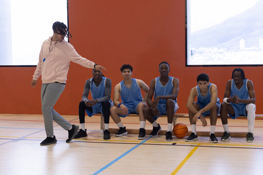Diverse male basketball team with coach directing players sitting on wooden bench in gym - Powered by Adobe