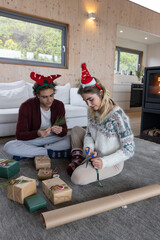 Naklejka premium Couple sitting on gray rug in living room wrapping gifts with scissors, ribbon and kraft paper