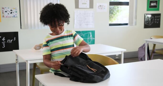 African boy entering classroom, unzipping black backpack on white desk pulling red folder for class
