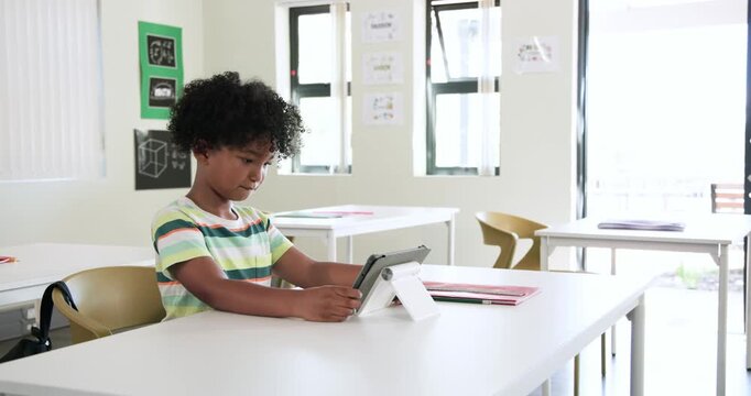 African American boy student starting tapping tablet on stand, sitting in classroom doing classwork