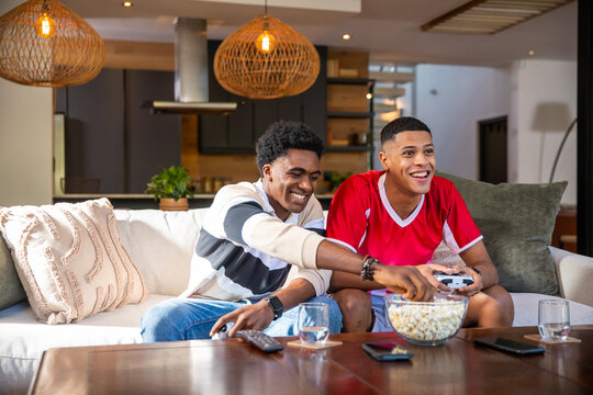 Diverse male friends holding controllers and snacking on popcorn bowl on sofa in modern living room