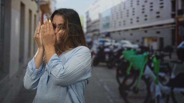 Woman covering face with hands on street near parked bicycles and building; shyness vulnerability privacy.
