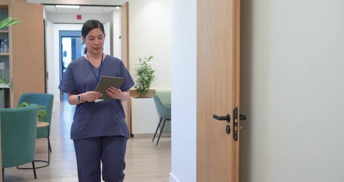 Woman nurse in scrubs entering clinic corridor tapping tablet reviewing chart and adjusting ID