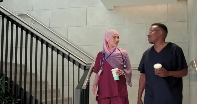 Diverse coworkers in scrubs descending stairs with coffee, apple and stethoscope, chatting on break