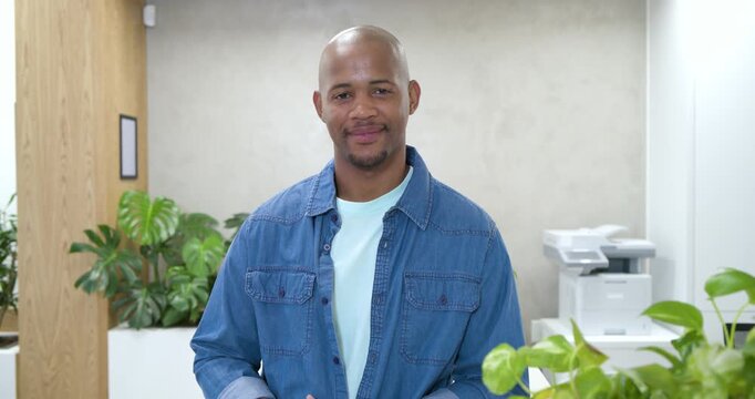 Adult African man shifting hands, leaning in, smiling as camera moving closer in office near plants