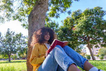 Young woman enjoying reading book in park, sitting on grass leaning against tree trunk, enjoying nature and relaxation © Javier