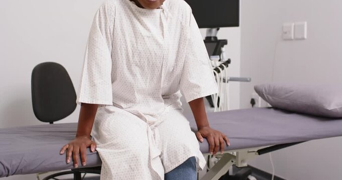 Middle-aged woman sitting on padded exam table in clinical room, preparing for exam, shifting gown