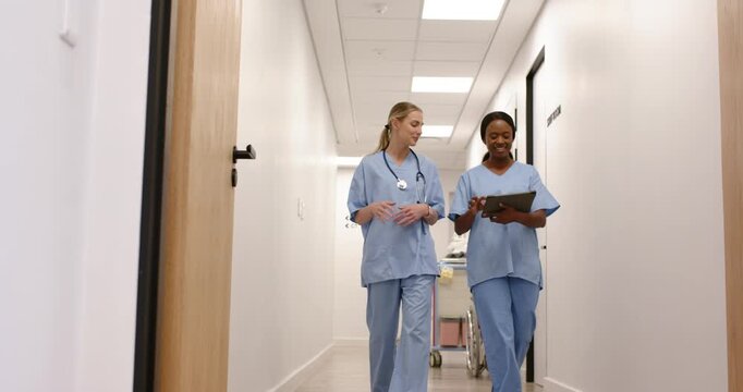 Diverse female RNs in scrubs walking hospital hall, right RN pointing tablet, planning care by cart