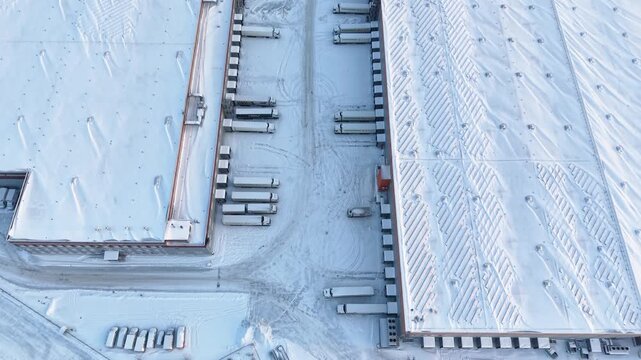 Drone video showcasing a snow-blanketed industrial warehouse complex. The aerial footage captures the expansive layout, showcasing parked trucks and a large, frosty roof under clear skies.