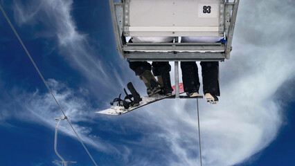 Snowboarders being transported to the ski slope. A section of the two-seater chairlift with passengers in the air. Feet in snowboard boots and snowboards with bindings.
