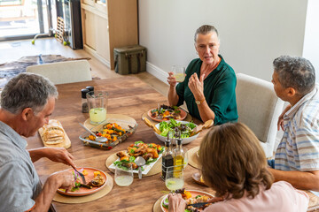 Diverse senior friends sharing midday meal on dining table at home with salad and lemonade