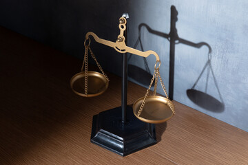 A scale of justice standing on a wooden surface against a textured grey wall. Legal balance,...