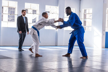 Diverse martial artists sparring while referee observing movements in dojo with belted gis, mats