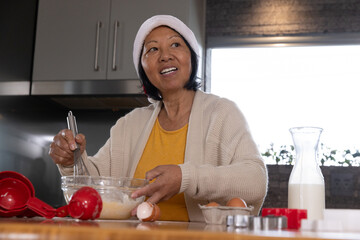 Senior Asian woman whisking batter at kitchen counter with glass bowl, red spoons, egg carton