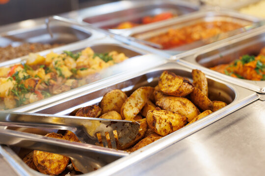 Potatoes in a buffet serving tray with tongs next to other dishes.