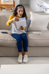 Teenage girl reading opened letter on sofa in home living room with smartphone, sealed envelope