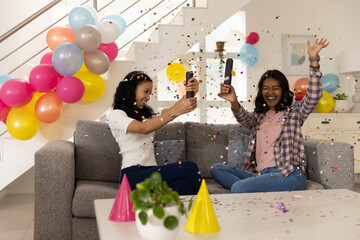 Obraz premium Diverse mom and teenage daughter popping confetti cannons on gray sofa in living room