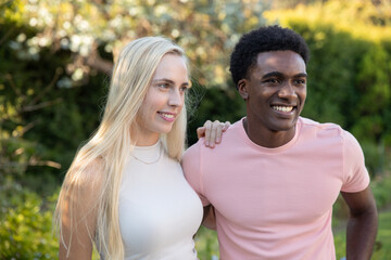 Diverse couple wearing sleeveless top and t-shirt standing in lush garden, smiling, gazing
