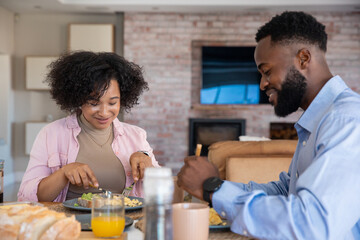 African american couple sitting at dining table cutting breakfast food, sipping orange juice
