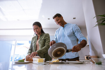 Diverse couple slicing bread and pouring eggs from frying pan at kitchen island with avocado