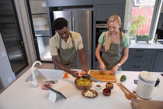 Diverse couple wearing aprons peeling carrots over bowl, slicing cucumbers on island in kitchen - Powered by Adobe