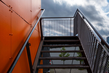 Modern staircase platform with orange mesh and railing under clouds using perspective and design to suggest upward progress and freedom