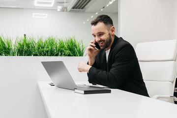 Smiling man in black suit is talking on a smartphone while sitting at a modern desk with a laptop and notebook, green plants visible in the background during a business meeting
