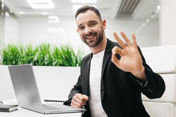 Smiling man in a black blazer is sitting at a desk with a laptop, making an okay gesture with his hand in a modern office environment with green plants