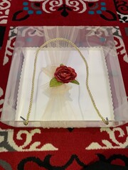 Clear box contains multiple small cakes  adorned with decorative element featuring red artificial flower and green leaves tied golden rope, suggesting wedding gift presentation.