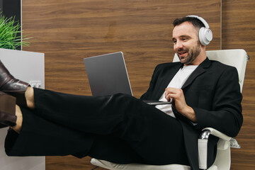 Male professional in a black suit and headphones is relaxing in a modern office chair with feet on a desk while using a laptop during a work break