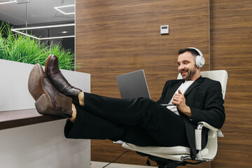 Businessman in a black suit and headphones is relaxing in an office chair with feet on a desk while using a laptop, modern decor and greenery visible in the background