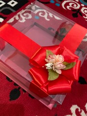 Clear box contains multiple small cakes  adorned with decorative element featuring red artificial flower and green leaves tied golden rope, suggesting wedding gift presentation.