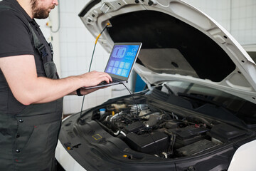 Caucasian young adult man using laptop computer while diagnosing engine in car repair shop, standing next to open hood and focusing on automotive diagnostics process