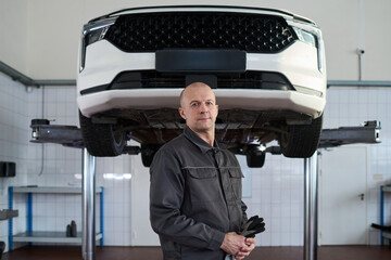 Portrait of middle aged Caucasian man standing in auto repair shop under elevated car, holding gloves and looking at camera, representing professional mechanic at workplace
