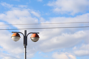 A street light with two spherical globes and orange caps stands against a bright blue sky filled with soft white clouds