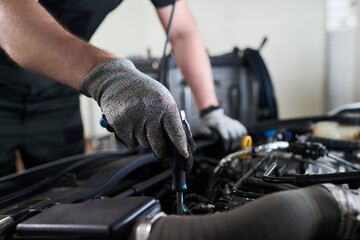 Man wearing work gloves repairing car engine in workshop, using screwdriver and focusing on mechanical components under open hood, demonstrating automotive maintenance skills