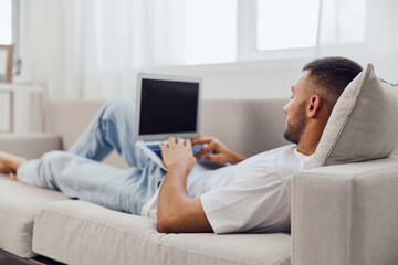 Relaxed man working on a laptop at home, wearing comfortable clothes and sitting on a cozy sofa, embodying the remote working lifestyle trend.