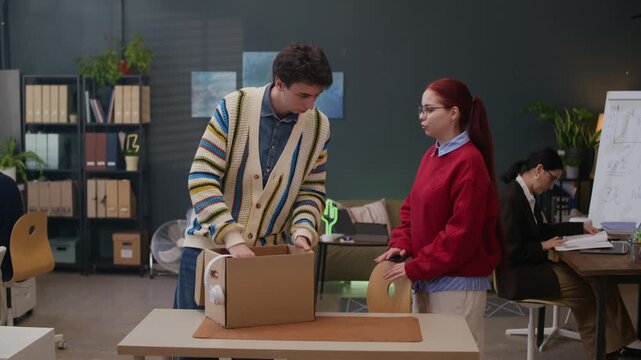 Long shot of young female manager onboarding male newcomer unpacking stuff from box in modern office workspace